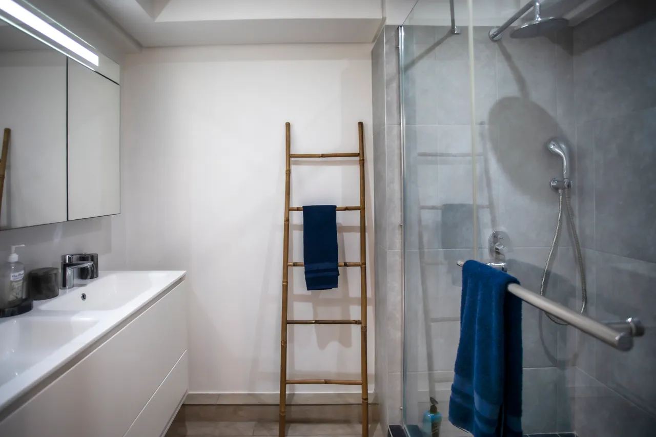 Salle de bain avec miroir large et évier moderne intégré.  
Douche à l'italienne avec parois en verre pour un accès pratique.  
Échelle en bois pour suspendre les serviettes à proximité.  
Éclairage LED au-dessus de l'évier assurant une bonne luminosité.  
Carrelage clair facilitant l'entretien quotidien de l'espace.