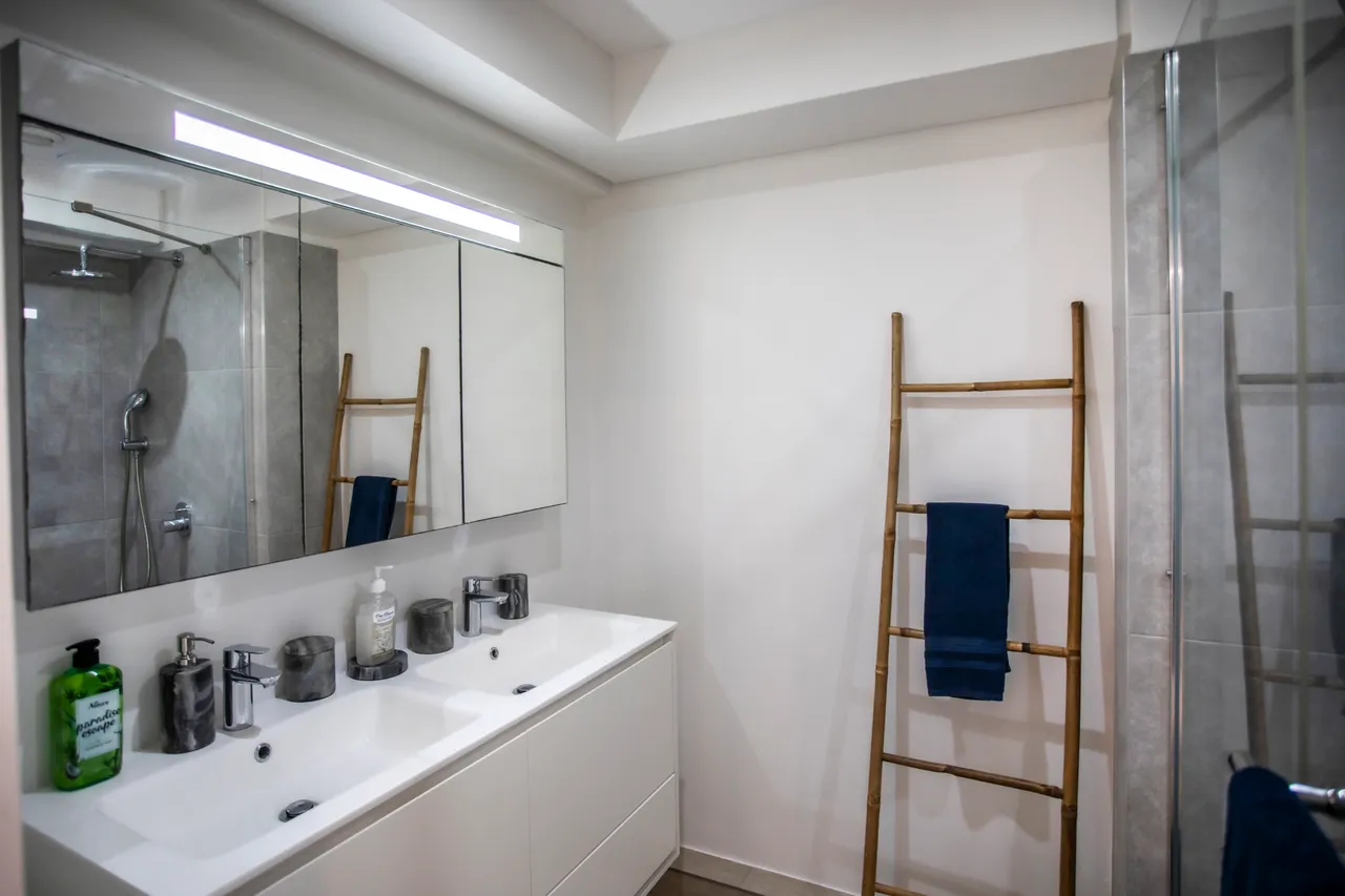 Salle de bain équipée de deux lavabos et d'un miroir large.  
Douche à l'italienne avec parois en verre pour un accès pratique.  
Échelle en bois pour suspendre les serviettes à proximité.  
Éclairage LED au-dessus des lavabos garantissant une bonne visibilité.  
Carrelage clair facilitant l'entretien quotidien de l'espace.