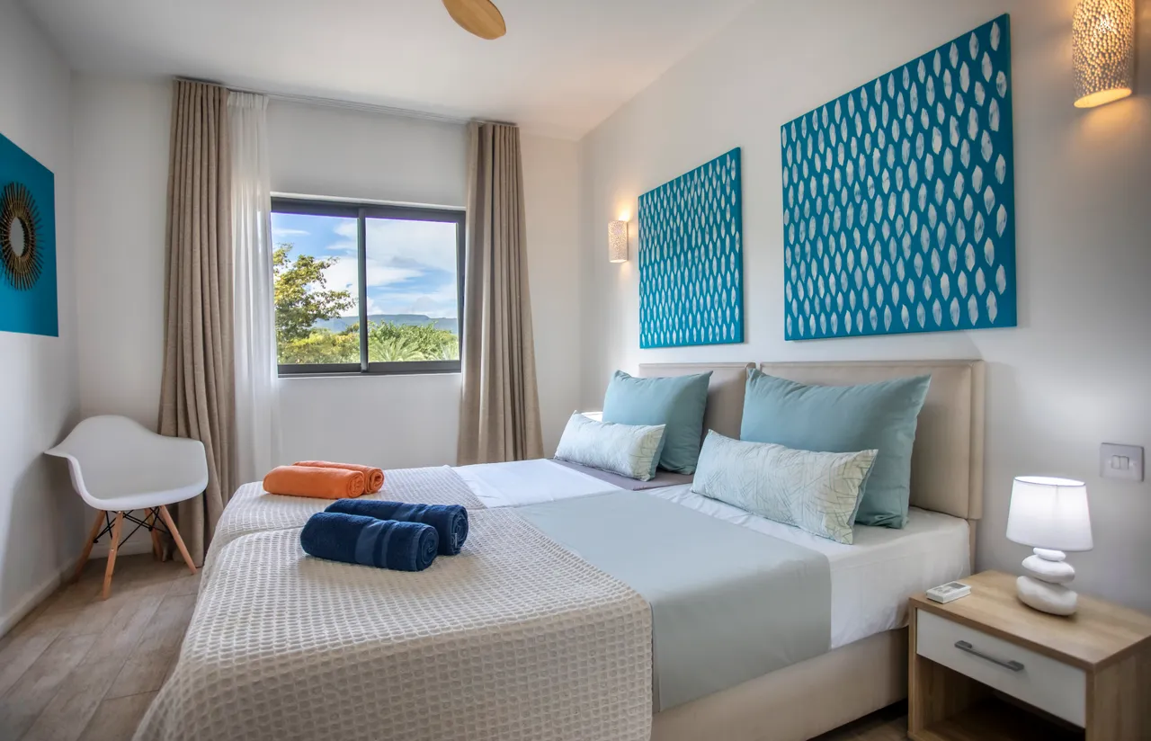 Chambre avec deux lits séparés et décoration murale colorée.  
Fenêtre large offrant une vue sur le jardin environnant.  
Éclairage mural et ventilateur de plafond pour un confort optimal.  
Draps coordonnés ajoutant une touche de couleur à l'espace.  
Sol en bois clair apportant une ambiance chaleureuse.