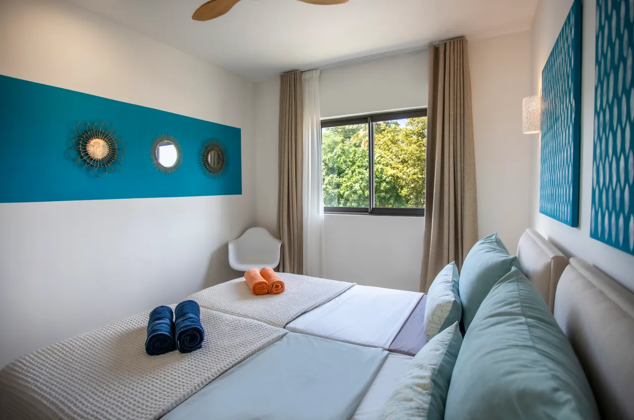 Chambre avec deux lits séparés et décor murale en bleu.  
Fenêtre offrant une vue sur le jardin verdoyant.  
Éclairage mural et ventilateur de plafond pour le confort.  
Tissus colorés ajoutant une note vive à l'espace.  
Sol en carrelage clair facilitant l'entretien quotidien.