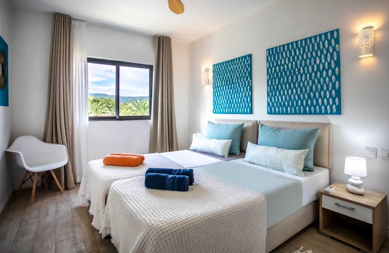 Couleurs apaisantes sur les murs et textiles de la chambre.  
Deux lits séparés avec linge de lit coordonné et coloré.  
Vue sur le jardin verdoyant grâce à la fenêtre spacieuse.  
Lumière naturelle et ventilateur de plafond pour le confort.  
Sol en bois clair ajoutant une touche chaleureuse à la pièce.