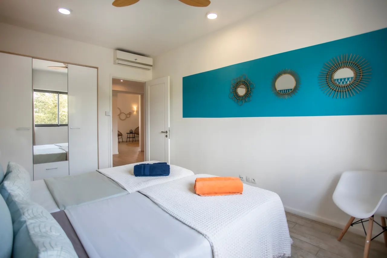 Chambre avec deux lits séparés et mur décoré de miroirs ronds.  
Fenêtre large offrant une vue sur l'extérieur verdoyant.  
Ventilateur de plafond et lumière naturelle pour un confort optimal.  
Draps en textile léger ajoutant une touche de couleur à l'espace.  
Sol en carrelage clair facilitant l'entretien quotidien.