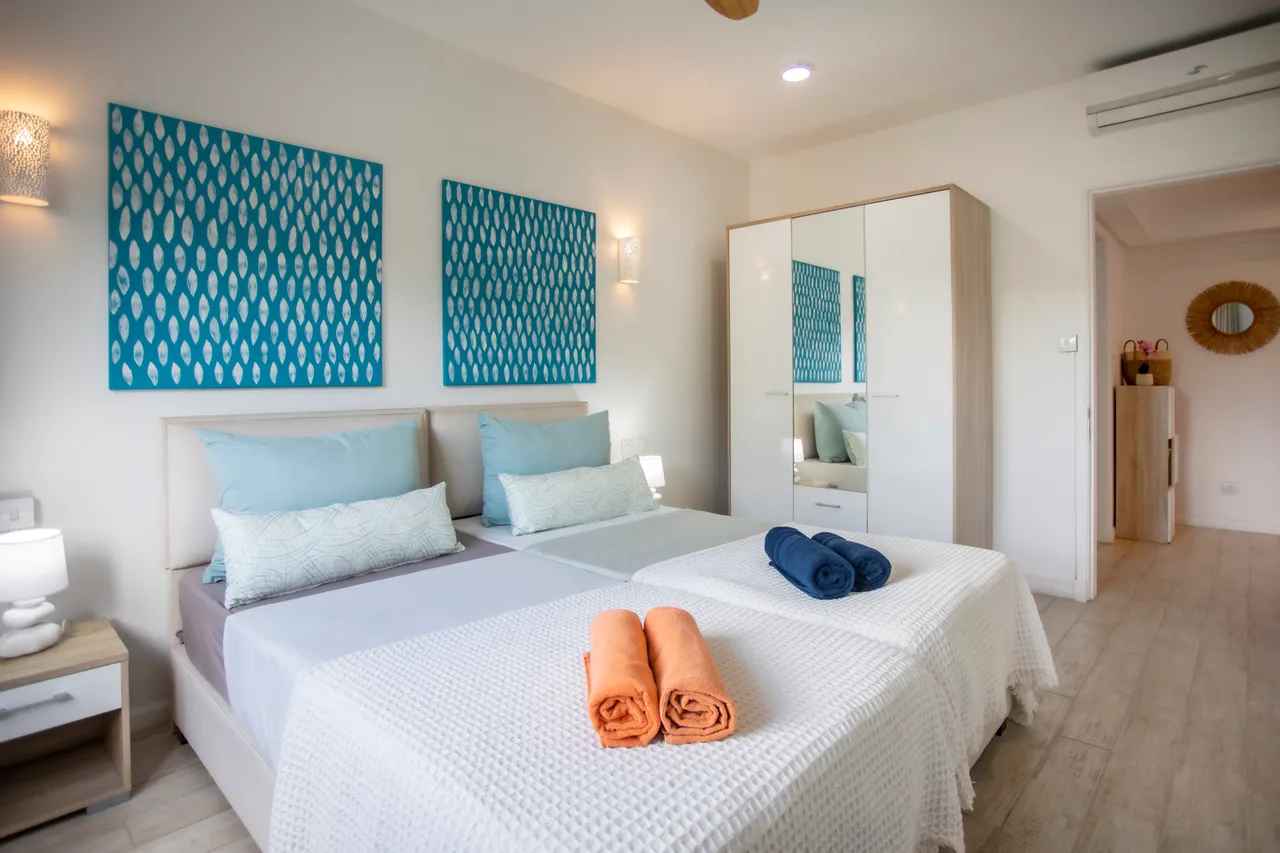 Chambre lumineuse avec deux lits séparés et textiles colorés.  
Fenêtre grande offrant une vue sur le jardin luxuriant.  
Ventilateur de plafond et éclairage doux pour un confort agréable.  
Rangement intégré avec miroir et table de nuit.  
Sol en carrelage clair pour un entretien facile.