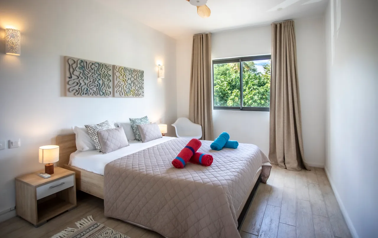 Chambre avec lit double et draps assortis de couleurs vives.  
Fenêtre large offrant une vue sur le jardin verdoyant.  
Lumière naturelle et éclairage d'appoint créant une ambiance agréable.  
Sol en bois clair ajoutant une touche chaleureuse à l'espace.  
Rideaux longs pour plus d'intimité et de confort visuel.
