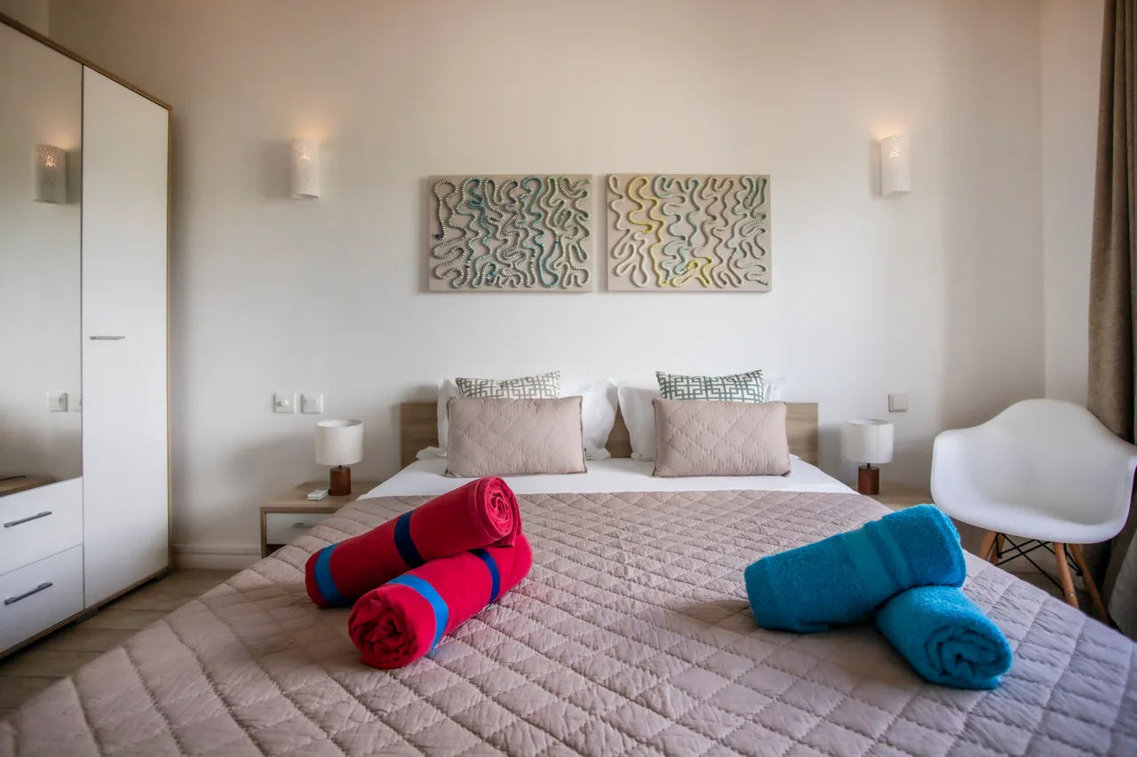 Chambre avec lit double, ornée de draps aux couleurs vives.  
Deux tableaux modernes ajoutent une touche artistique au mur.  
Éclairage doux et lampes de chevet pour une ambiance apaisante.  
Rangement intégré avec armoire offrant un espace pratique.  
Fenêtre laissant entrer la lumière naturelle, créant une atmosphère lumineuse.