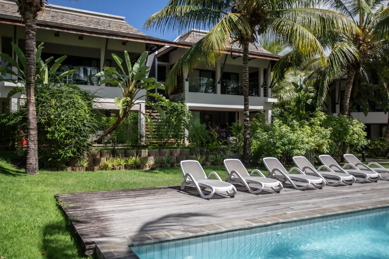 Terrasse en bois devant la piscine, entourée de chaises longues.  
Jardin verdoyant ajoutant une touche naturelle à l'espace extérieur.  
Palmiers offrant de l'ombre et une ambiance tropicale aux abords de la piscine.  
Accès direct à la piscine depuis la terrasse pour plus de commodité.  
Espace dégagé permettant de profiter du soleil en toute tranquill