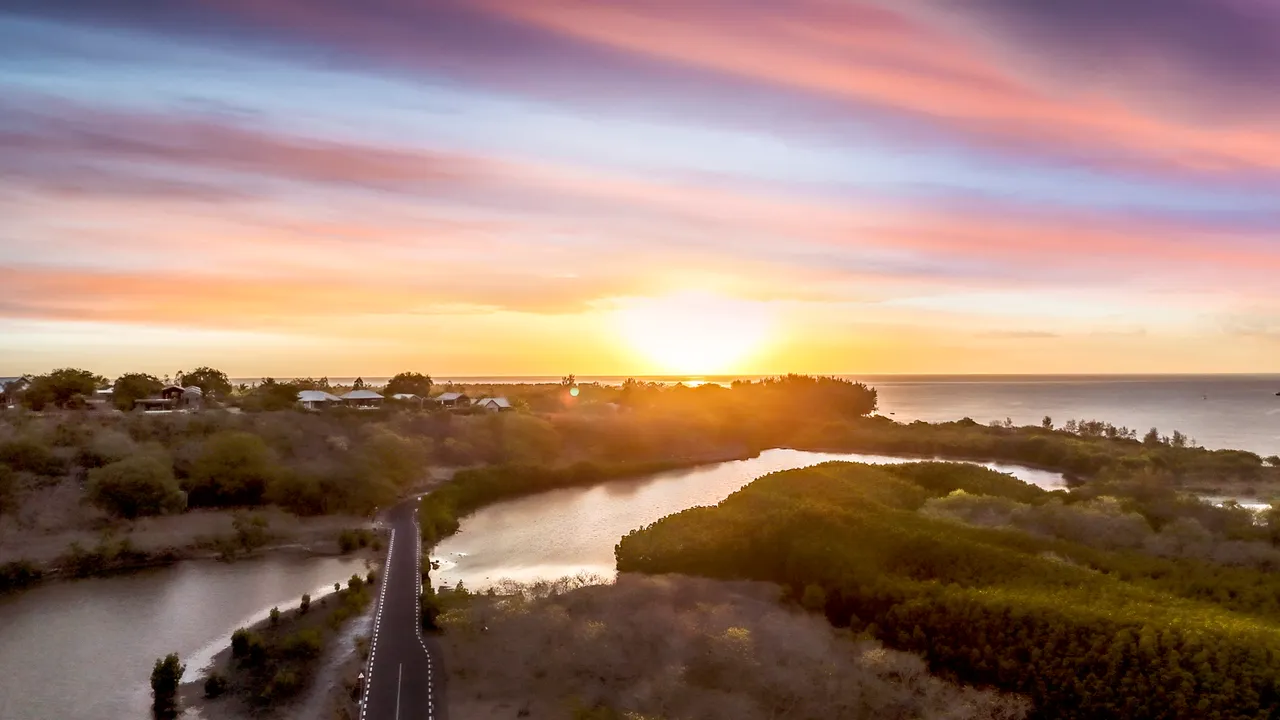 Coucher de soleil sur la mer, illuminant les collines verdoyantes environnantes. Route sinueuse bordée d'eau au premier plan.
