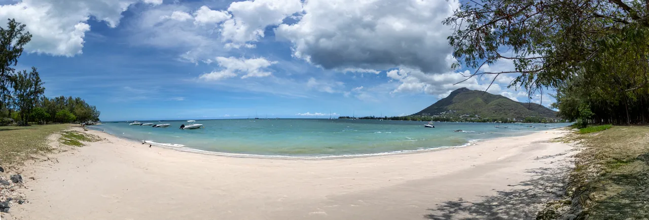 Plage de sable clair avec des eaux calmes et peu profondes. Vue sur la montagne à proximité. Espace vert avec des arbres en bordure de plage. Bateaux amarrés au large, ajoutant une touche maritime.