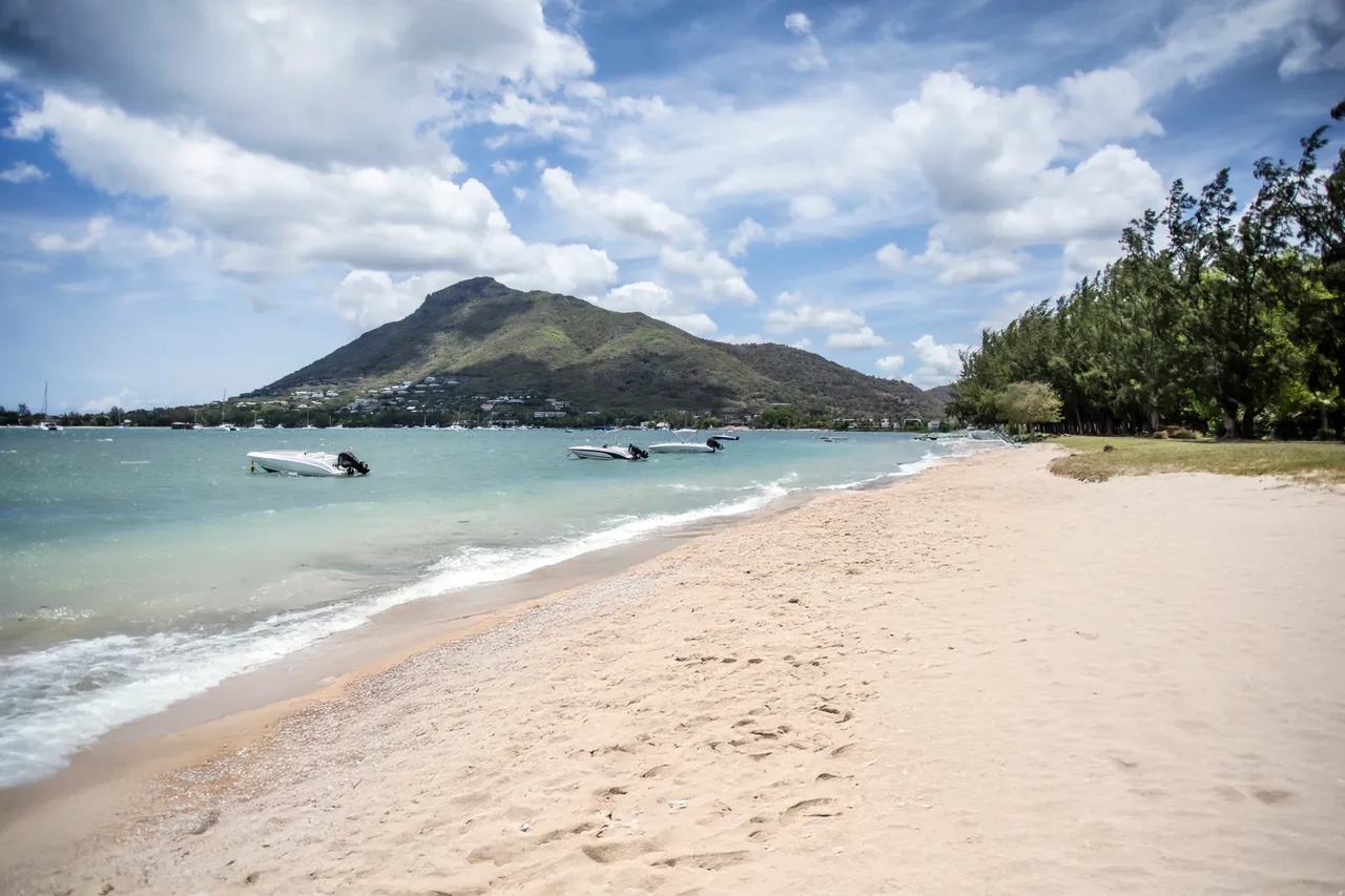 Plage de sable fin avec des bateaux amarrés au loin. Vue dégagée sur la montagne.