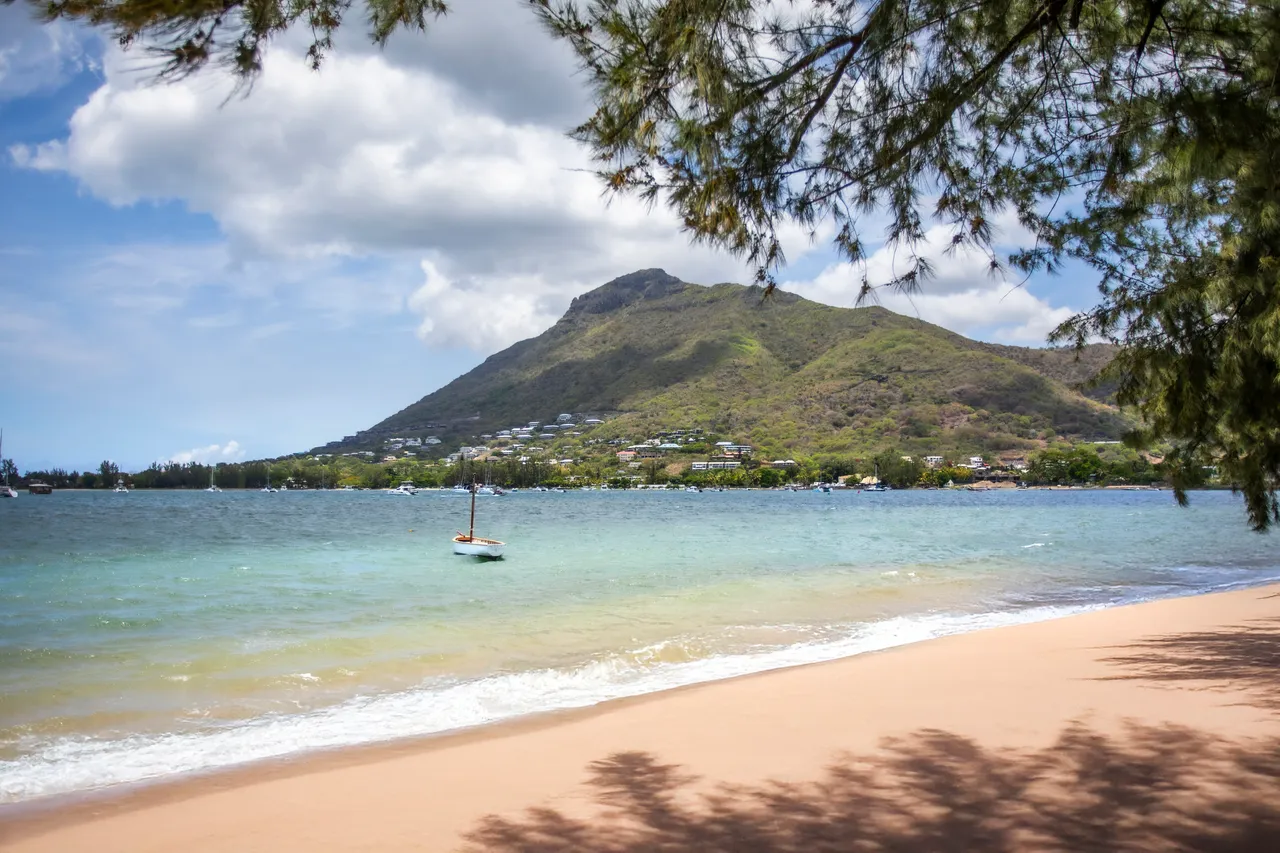 Plage de sable doux avec des eaux claires et un fond marin visible. Montagne verdoyante en arrière-plan, ajoutant de la profondeur à la vue.