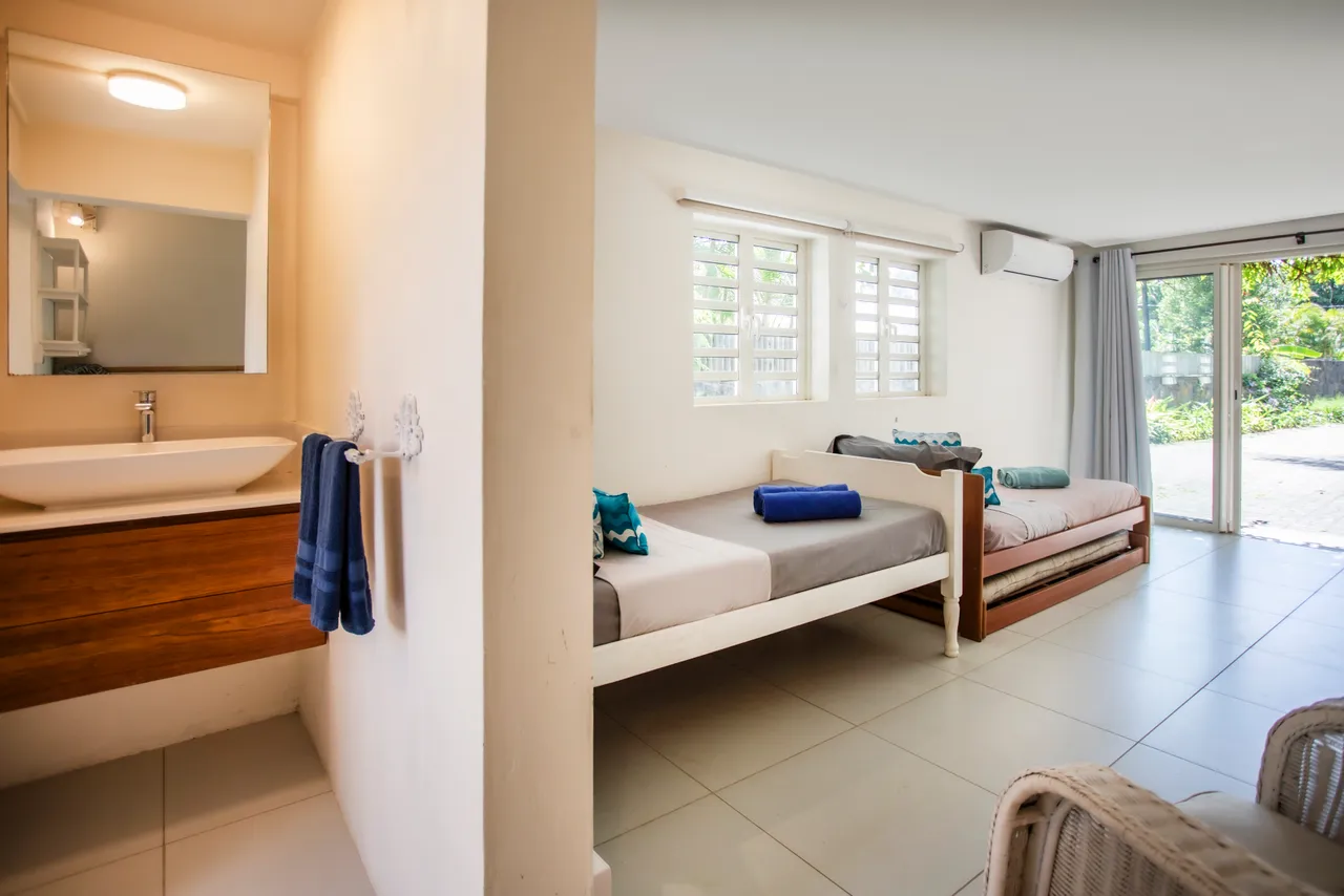 Salle de bain lumineuse avec lavabo moderne et miroir. Espace simple et fonctionnel, séparé par un mur. Chambre adjacente avec lit individuel et accès au jardin.