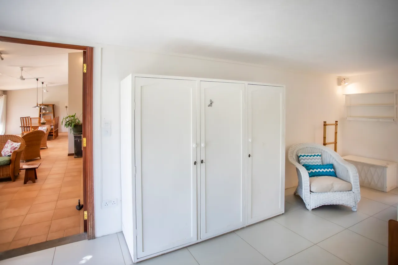 Espace de rangement blanc avec mobilier en rotin et accès à la salle à manger. La lumière naturelle pénètre par l'entrée ouverte. Mobilier sobre et fonctionnel dans un agencement lumineux. Coin salon avec chaises en rotin et vue sur l'espace commun.