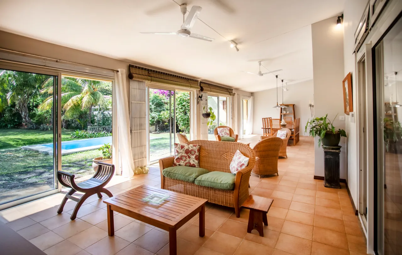 Sols en carrelage clair et mobilier en rotin dans la galerie. Espace lumineux avec de grandes baies vitrées. Coin salon avec chaises confortables et accès direct au jardin. Plantes tropicales visibles depuis l'intérieur, apportant une touche de verdure. Vue sur la piscine depuis l'espace de vie.