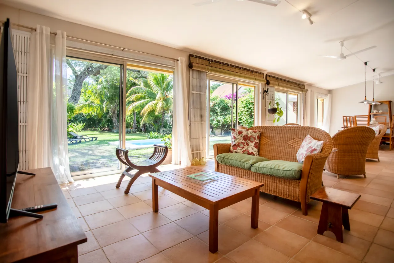 Lumière naturelle abondante grâce aux grandes baies vitrées. Coin salon avec mobilier en rotin et tables basses en bois. Vue directe sur le jardin et la piscine depuis l'intérieur. Espace agréable avec plantes tropicales en arrière-plan.