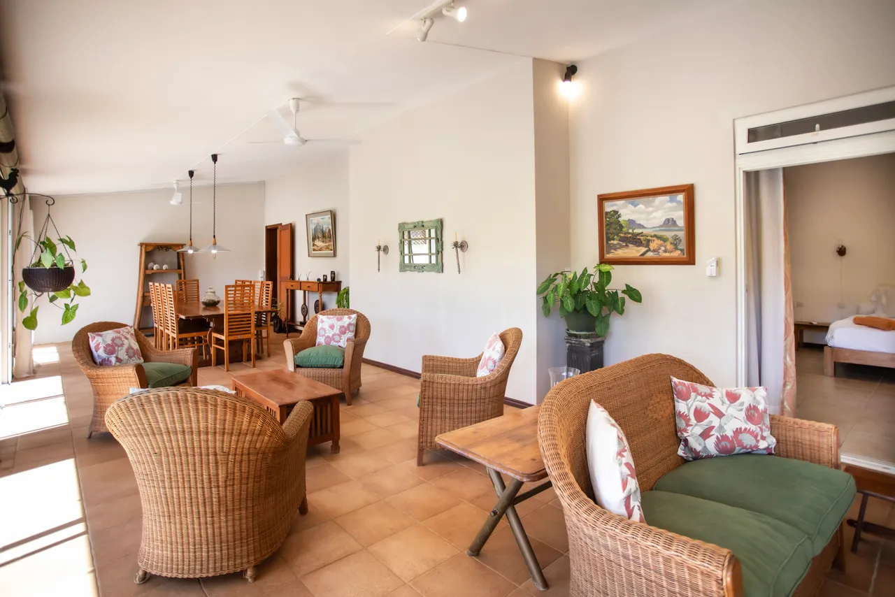 Galerie avec mobilier en rotin et tables en bois. Espace bien éclairé avec vue sur le jardin. Salle à manger attenante avec chaises en bois et décor simple.