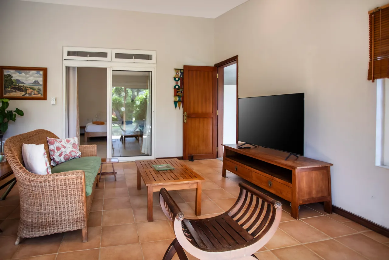 Sols en terre cuite et mobilier en rotin dans un espace de vie lumineux. Vue sur le jardin depuis le canapé. Télévision et accès direct à la chambre.