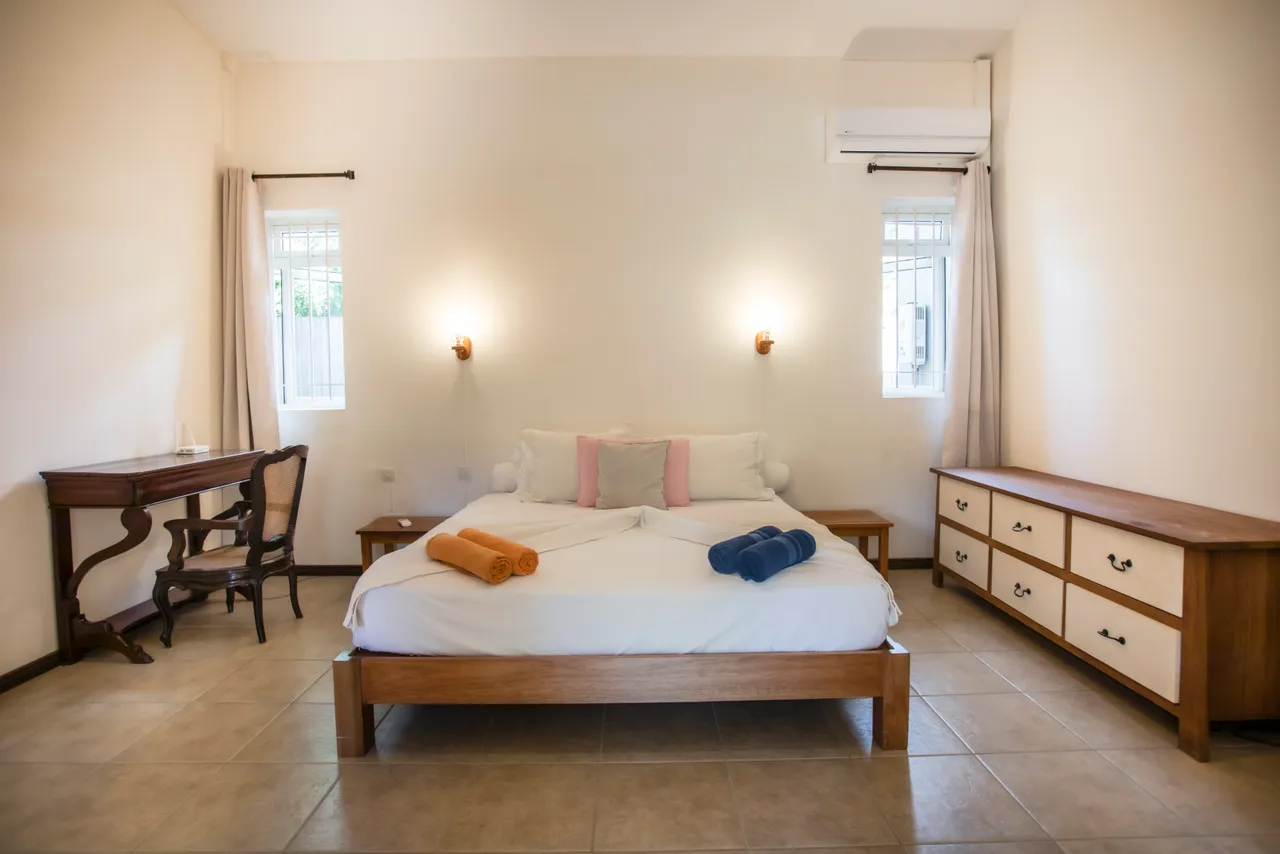Chambre avec lit double, bureau en bois et fenêtres apportant lumière naturelle. Espace de rangement disponible et sols en carrelage.