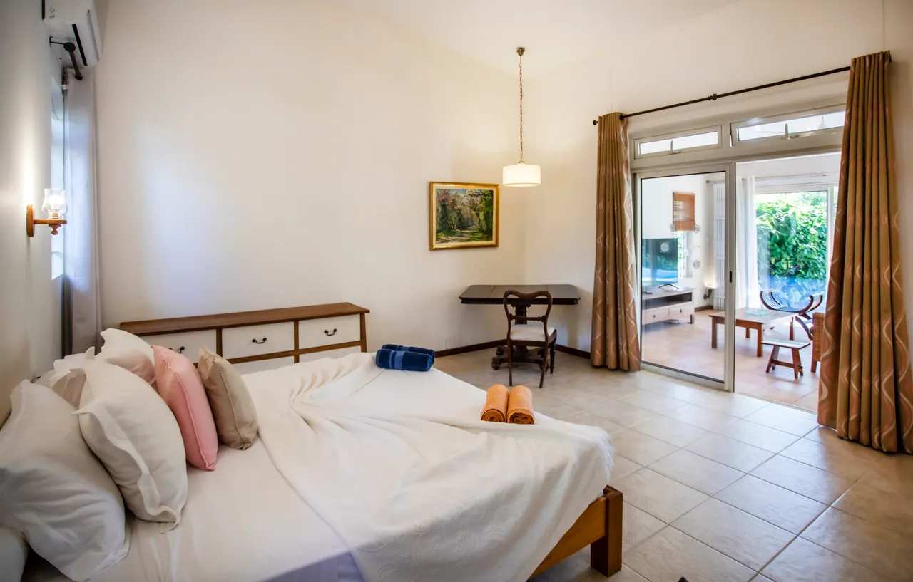 Chambre avec un sol en carrelage et literie blanche. Bureau en bois et accès direct au jardin. Fenêtres panoramiques offrant une vue sur l'extérieur.