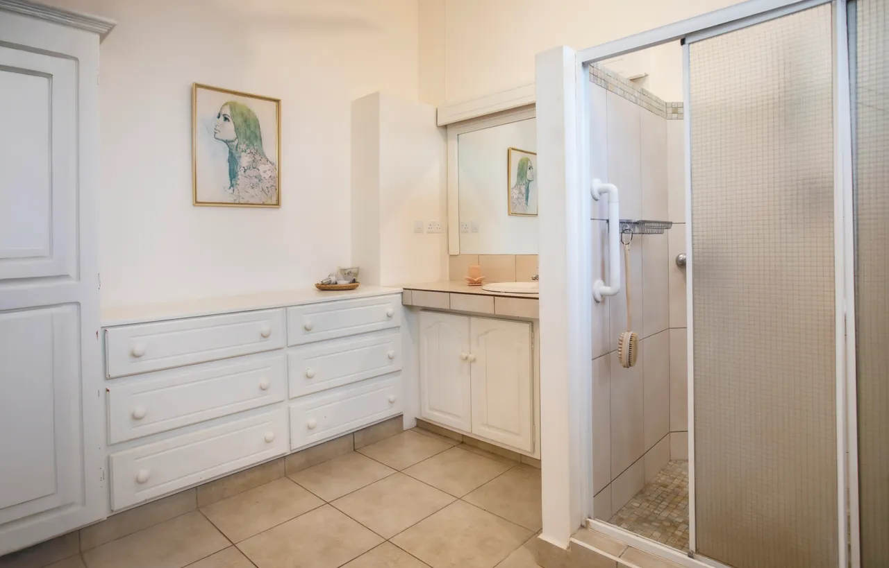 Sols en carrelage clair et meuble de rangement dans la salle de bain. Douche spacieuse et éclairage naturel par fenêtre.