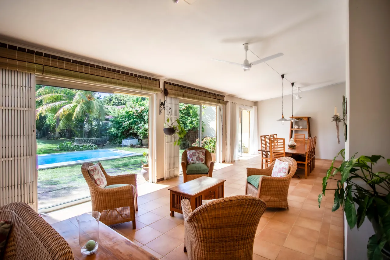 Sol en carrelage clair avec mobilier en rotin et coussins colorés. Espace ouvert avec de grandes baies vitrées donnant sur la piscine. Coin repas en bois exposé à la lumière naturelle. Ventilateurs de plafond pour une circulation d'air agréable.