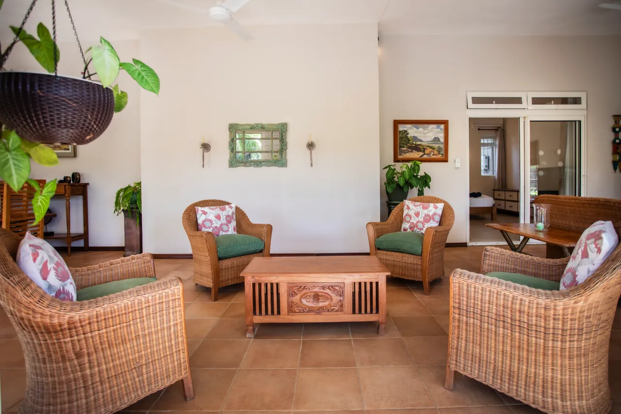 Teintes naturelles des fauteuils en rotin, créant un espace accueillant. Espace ouvert avec accès à la terrasse et au jardin. Table en bois au centre, ajoutant une fonctionnalité pratique. Plantes vertes apportent une touche de vie à la galerie.