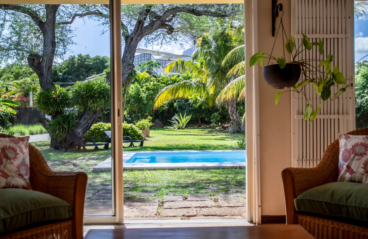 Sols en pierre et grandes baies vitrées offrant une vue sur le jardin. Accès direct à la piscine depuis la galerie. Fauteuils en rotin ajoutent une touche de confort à l'espace. Plantes vertes encadrent la vue sur l'extérieur, apportant une touche de nature.