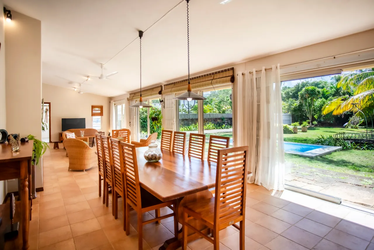 Galerie spacieuse avec une grande table en bois et des chaises assorties. Éclairée par des baies vitrées offrant une vue sur le jardin. Accès direct à l'espace extérieur et à la piscine. Plantes vertes ajoutent une touche naturelle à l'environnement.