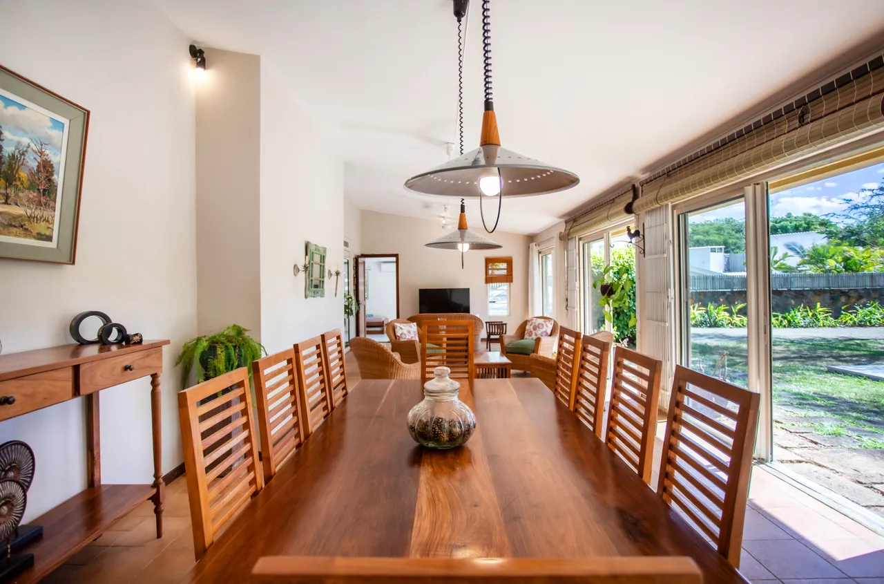 Salle à manger avec une grande table en bois et chaises en bois. Éclairée par des luminaires suspendus et baies vitrées. Accès direct au jardin et à la piscine. Mobilier au design simple et fonctionnel.
