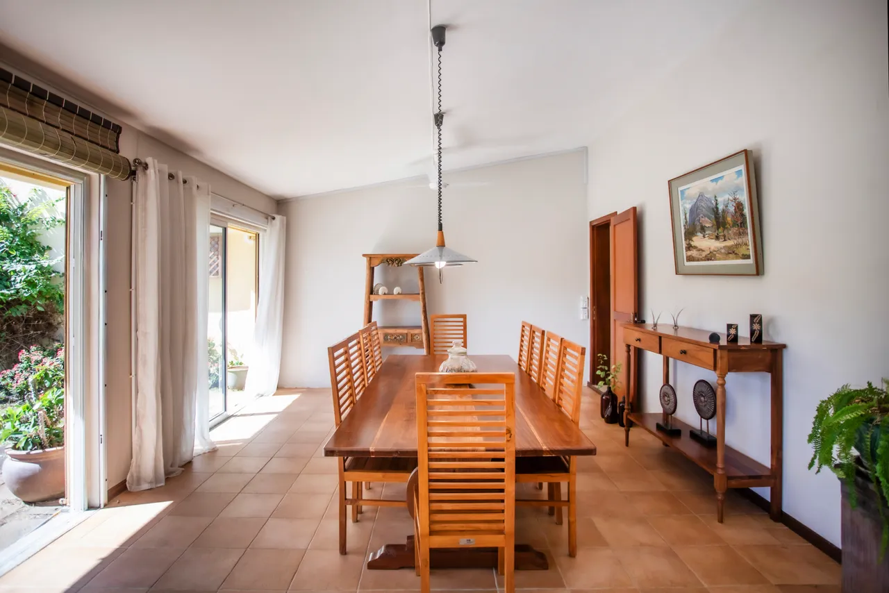 Espace repas lumineux avec baies vitrées ouvrant sur le jardin. Grande table en bois occupant le centre de la pièce. Éclairé par un luminaire suspendu, créant une ambiance conviviale.