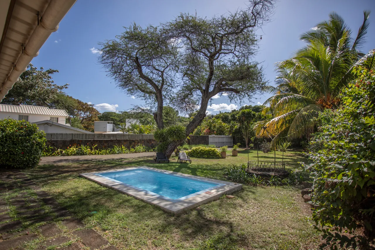 La piscine est entourée d'un jardin verdoyant, offrant une ambiance tranquille. Un grand arbre crée de l'ombre près de l'eau. Le sol en gazon invite à la détente à l'extérieur. Des espaces de repos sont aménagés autour de la piscine.