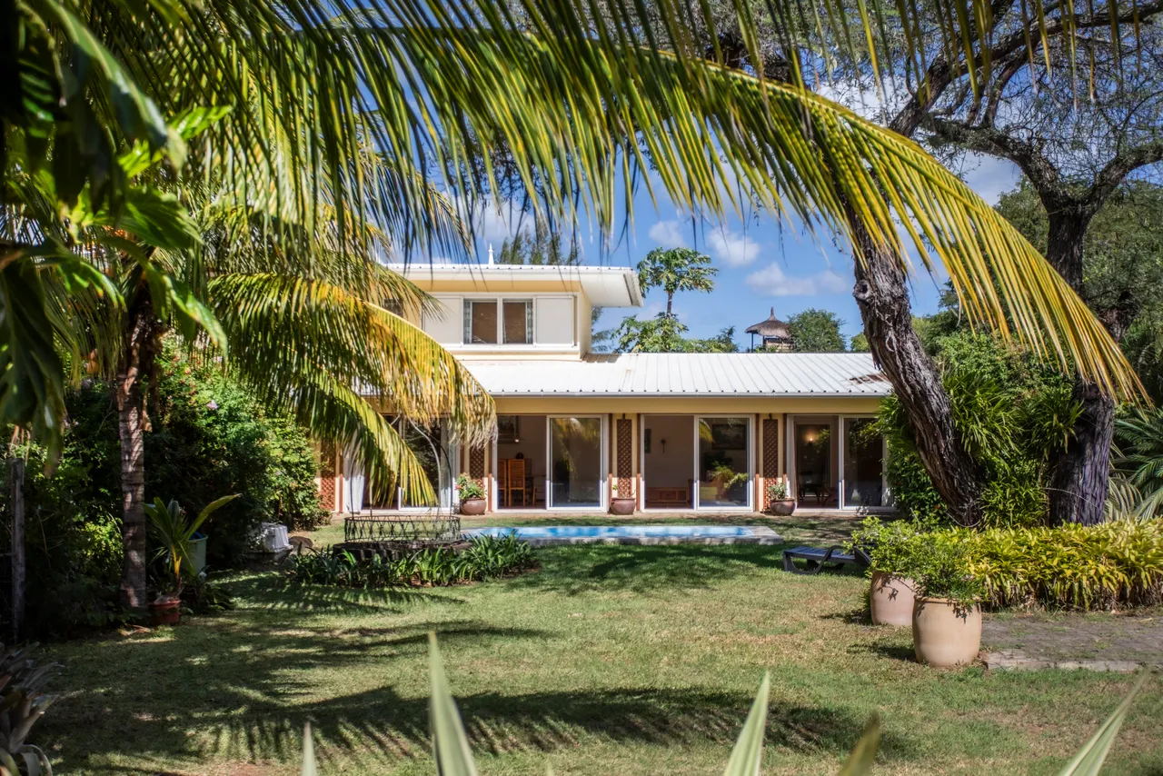 Bungalow avec piscine et accès direct à la plage, entouré de verdure. Les grandes baies vitrées apportent une lumière naturelle. Palmiers et plantes tropicales ajoutent une touche de verdure au jardin.
