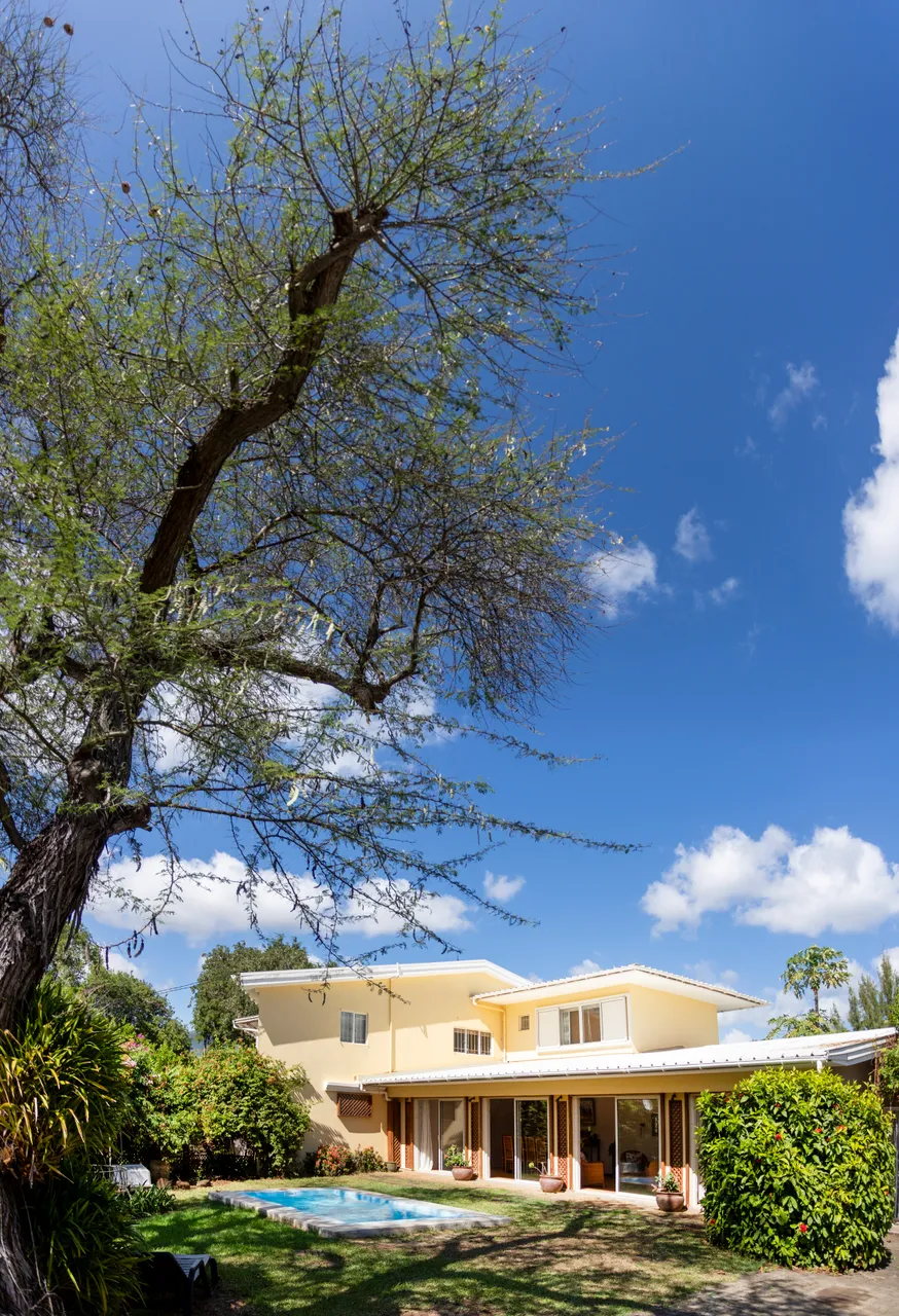 Vue du bungalow depuis le jardin, avec une piscine en arrière-plan. Arbre mature et verdure environnante apportent une atmosphère naturelle. Ciel dégagé et lumineux complètent l'environnement.
