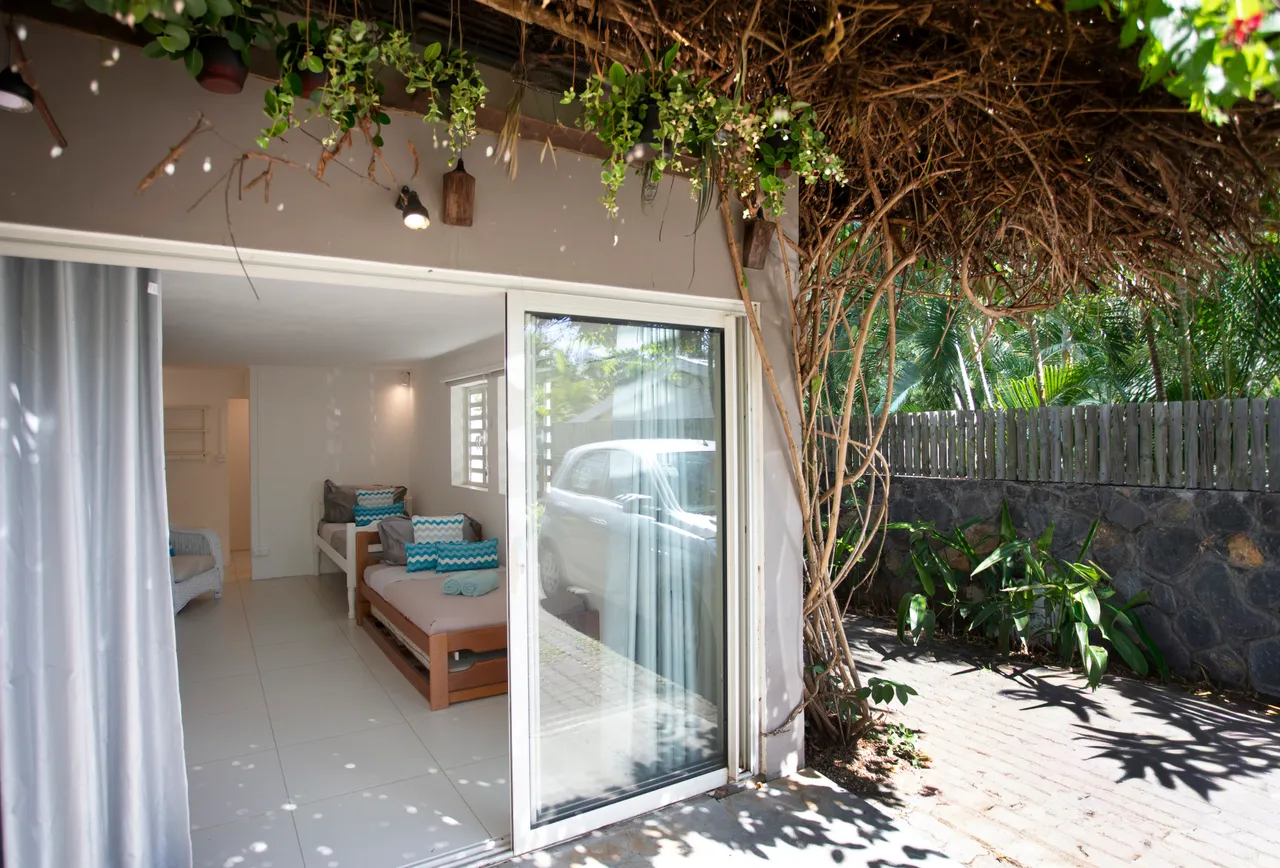 Sol carrelé dans un espace ouvert, avec un canapé et des coussins colorés. L'accès à l'extérieur est facilité par une grande baie vitrée. Verdure et plantes entourent l'entrée, créant une ambiance naturelle.
