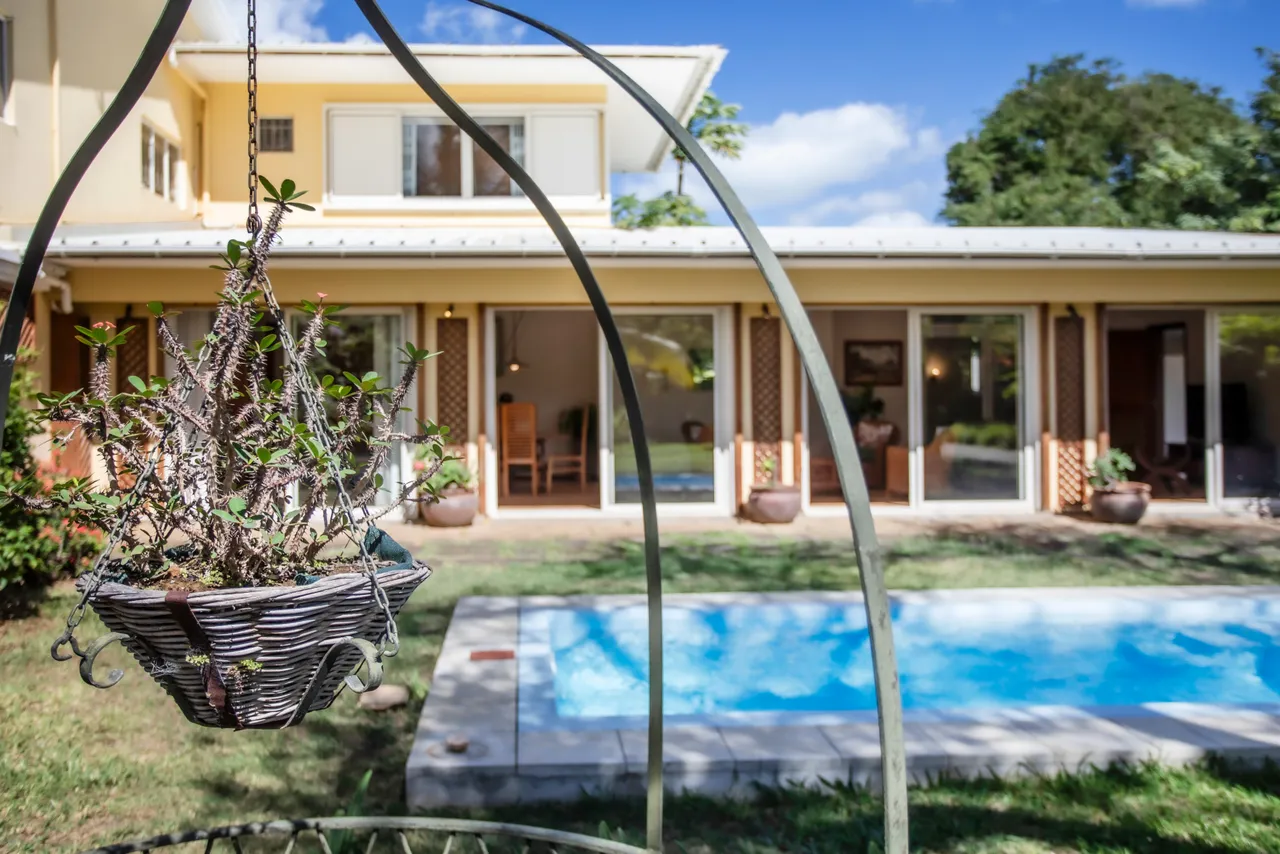 Lumière abondante dans le bungalow grâce aux grandes baies vitrées. La piscine est visible depuis le jardin, offrant un cadre relaxant. Un pot de fleurs suspendu ajoute une touche décorative à l'extérieur.