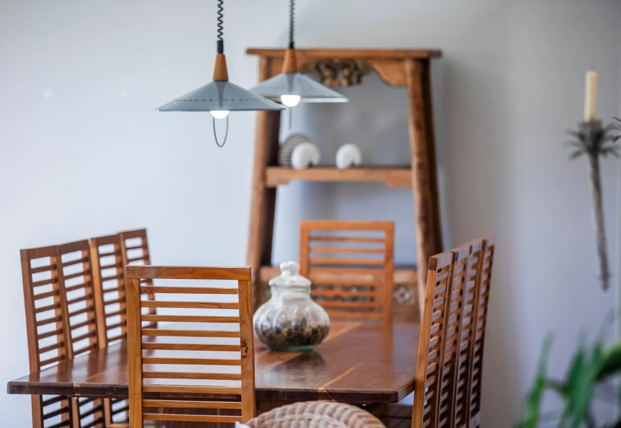 Table en bois avec chaises en lattes, située dans une galerie lumineuse. Éclairage suspendu au-dessus crée une ambiance agréable.
